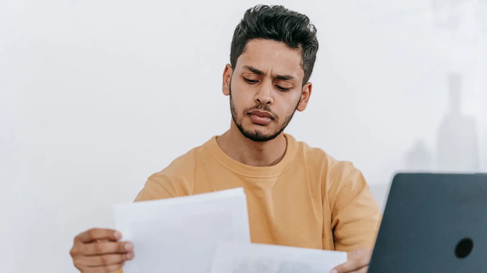 Man in a mustard T-shirt frowning slightly as he reads printed pages beside an open laptop