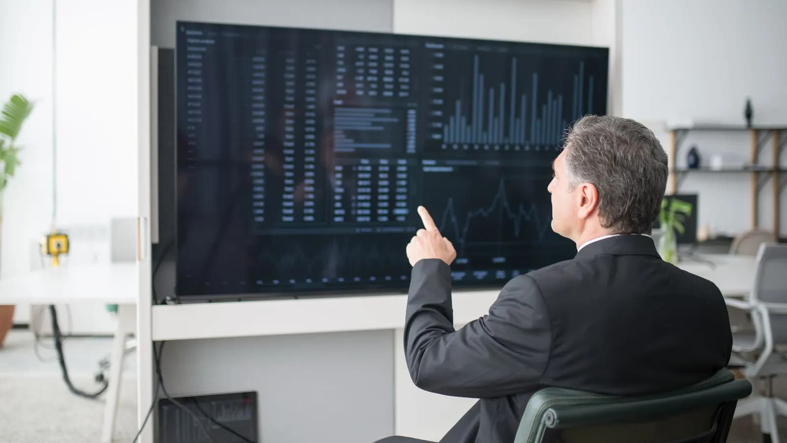 A man in a dark suit points at bar charts and tables on a large wall-mounted screen.