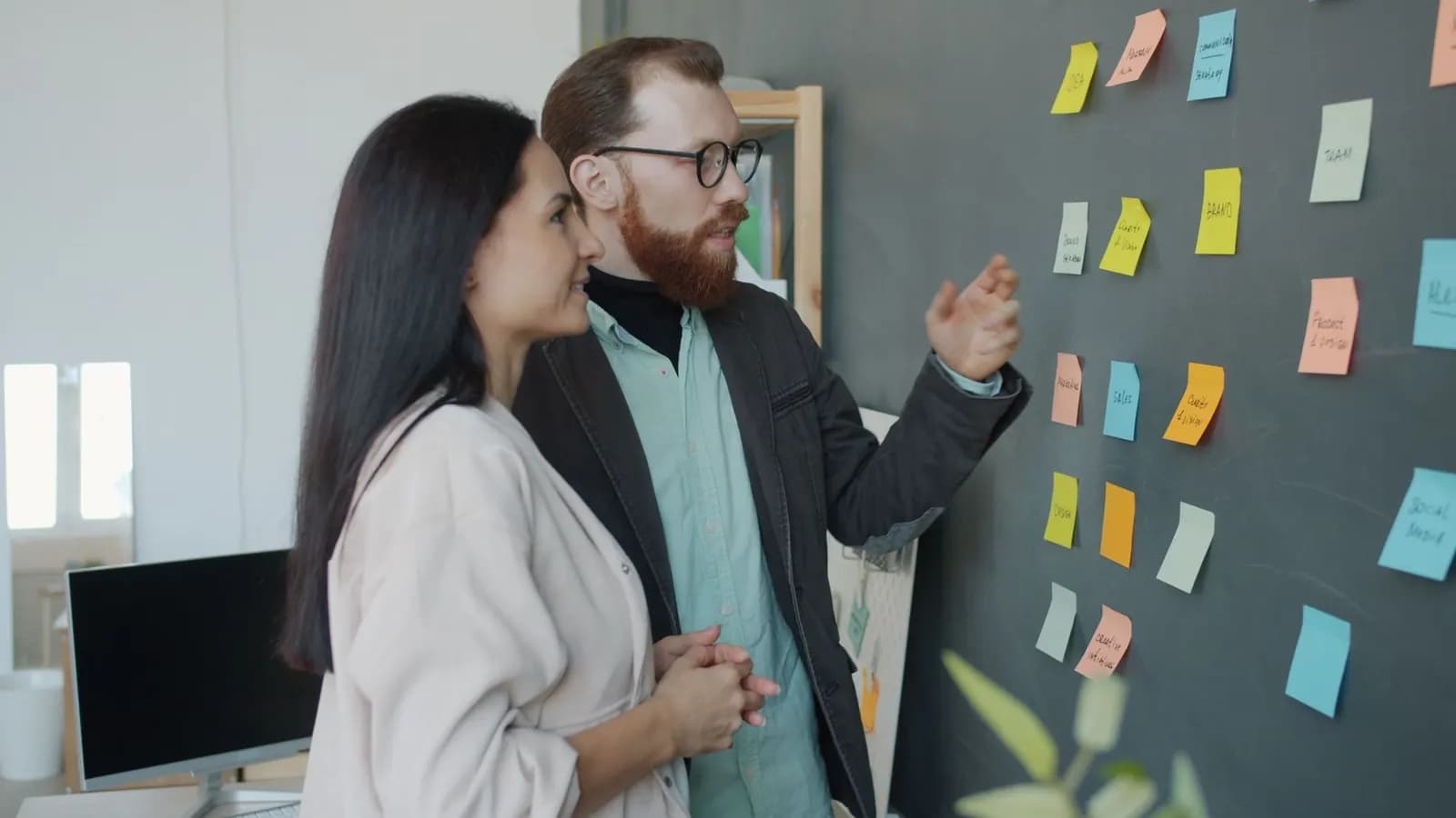 Two colleagues discuss colorful sticky notes arranged on a dark wall board in a bright office.