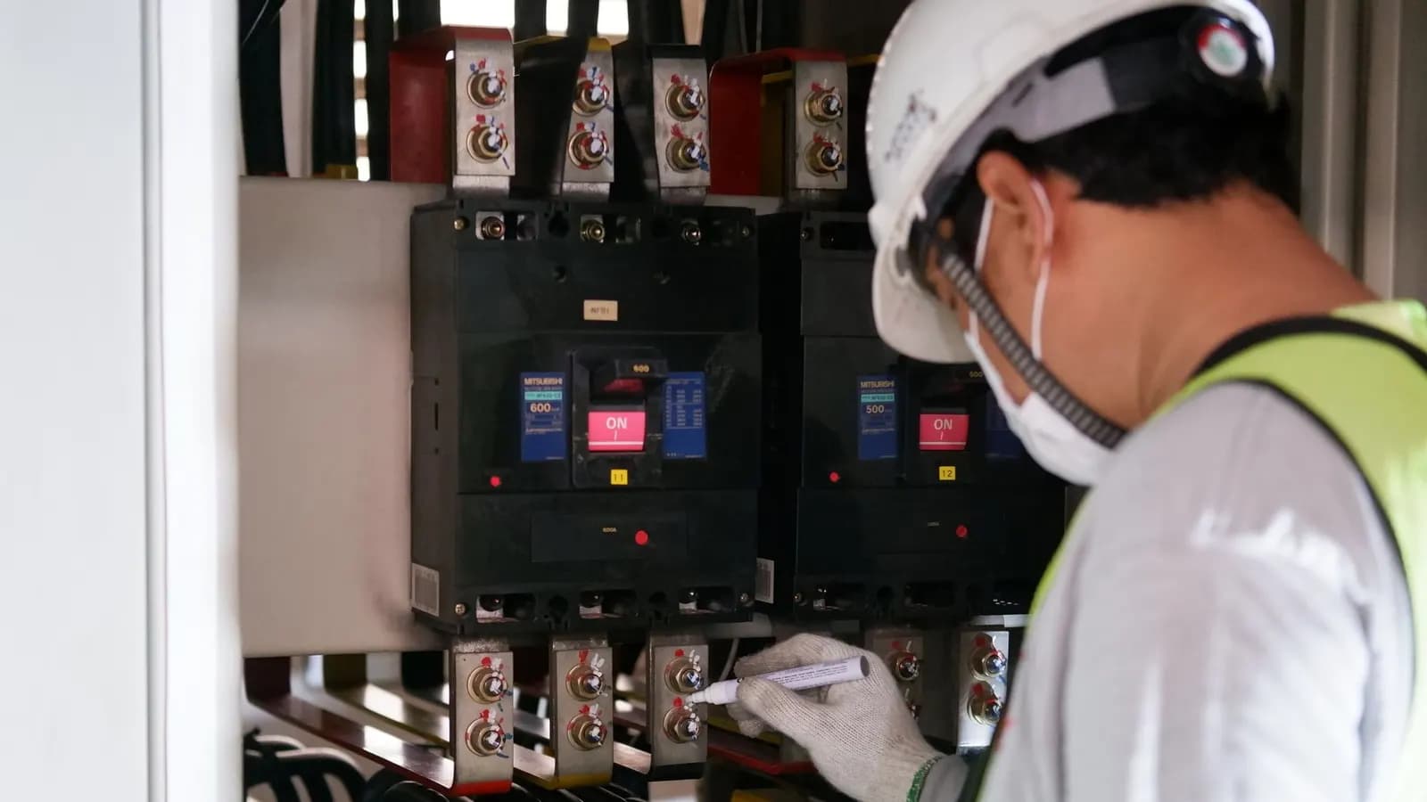 Technician in a hard hat and face mask checking a breaker panel with a test pen
