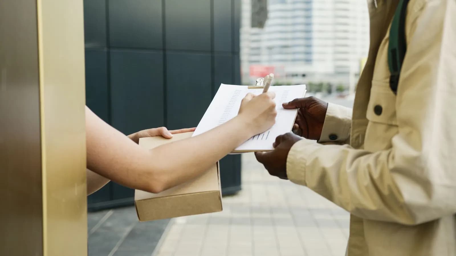 A person holding a small parcel signs a delivery receipt on a courier's clipboard.