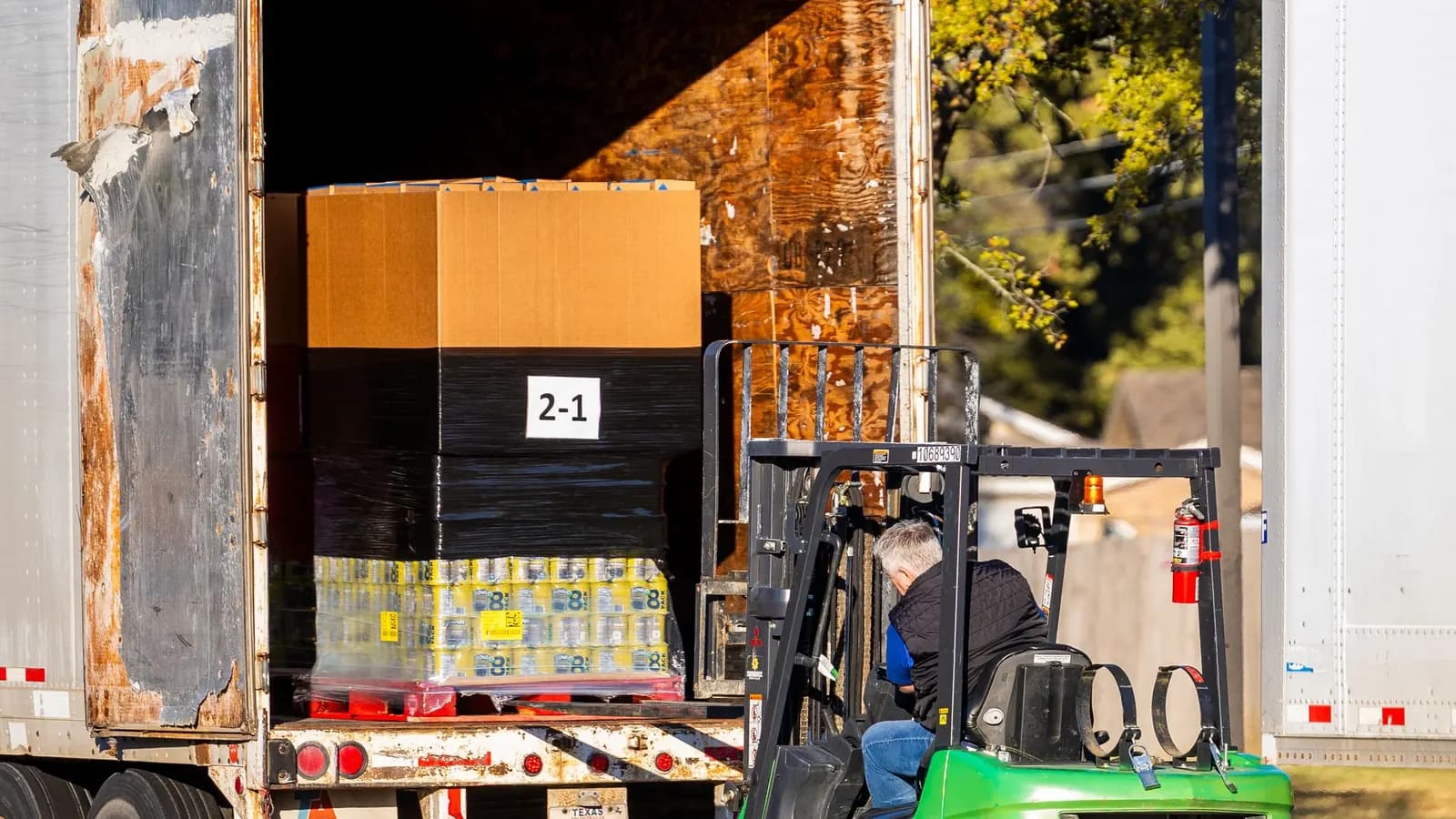 A forklift operator loads a shrink-wrapped pallet into an open freight trailer parked outdoors.