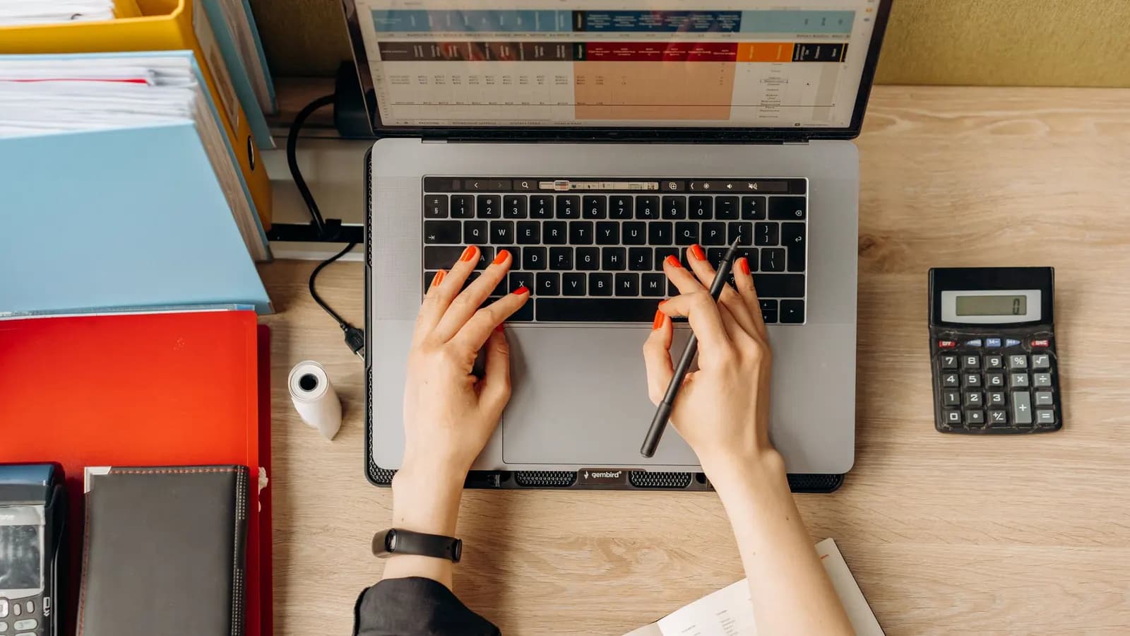 Overhead view of hands typing on a laptop beside a calculator, a pen and stacked office folders