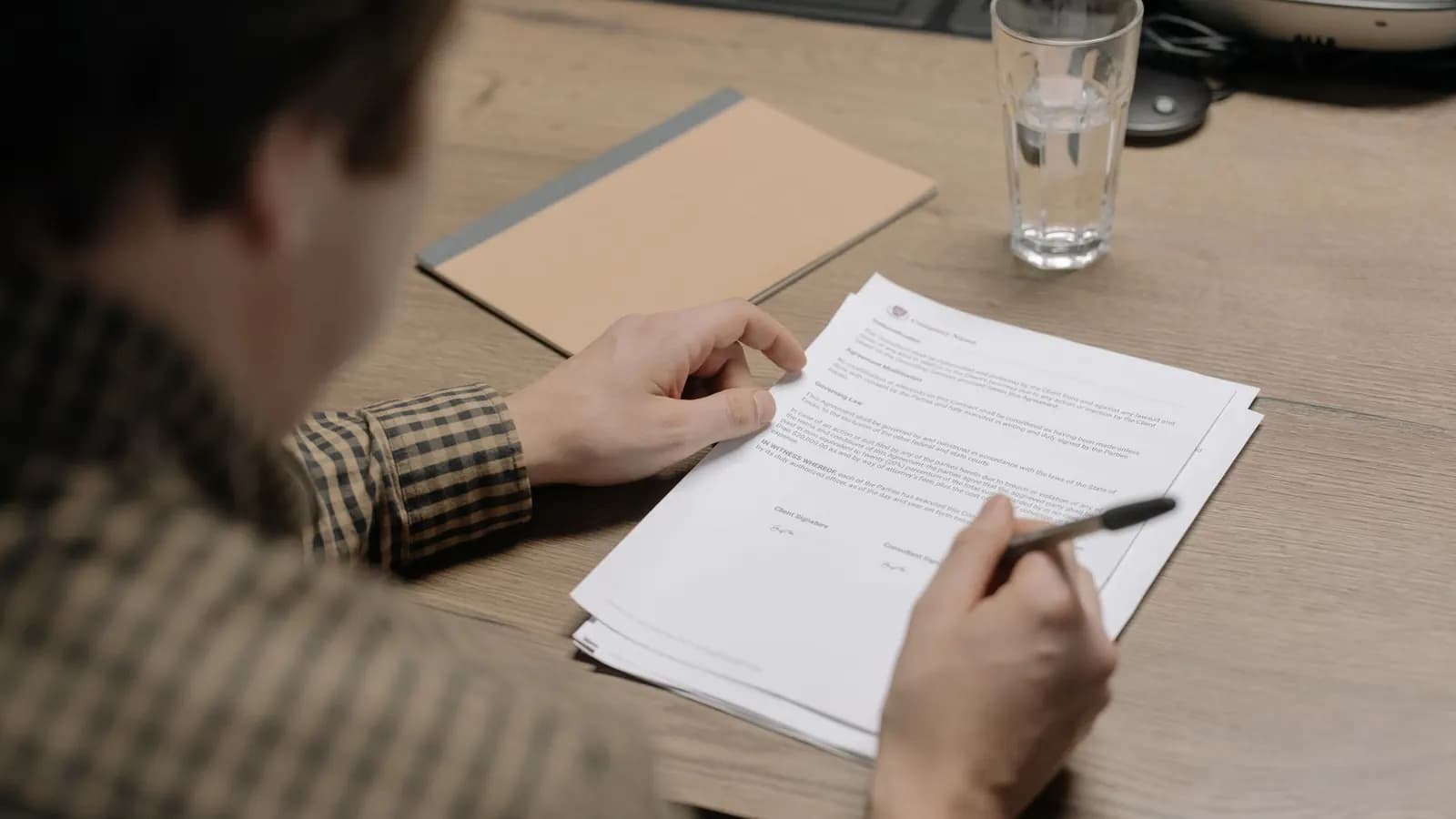 Man in a checked shirt reviewing a printed agreement with a pen at a wooden table