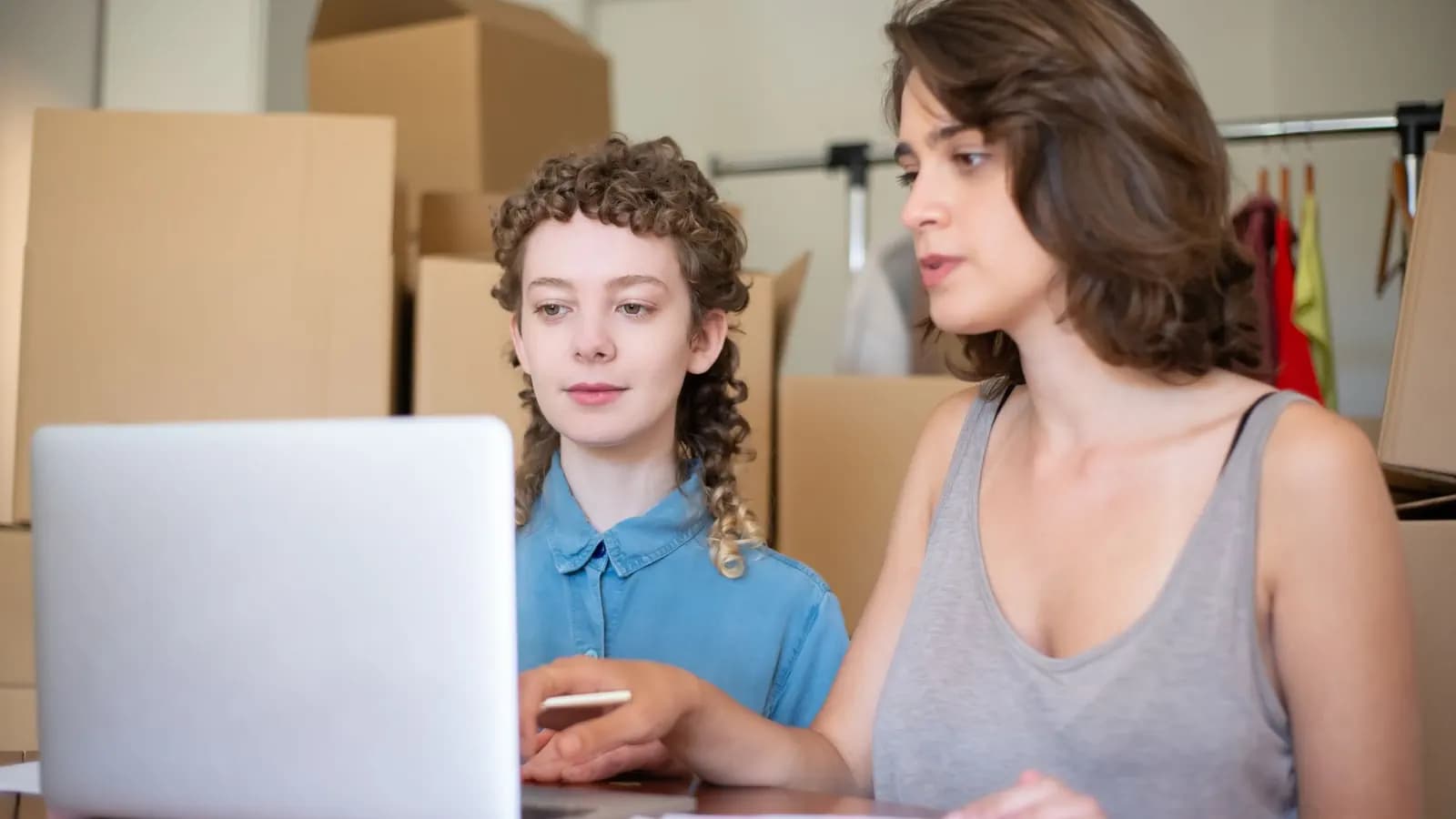 Two women talking over a laptop at a table, with stacked cardboard boxes and a clothing rack behind them