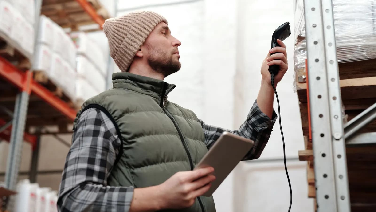 A warehouse worker in a beanie scans a shrink-wrapped pallet on a rack while holding a tablet.