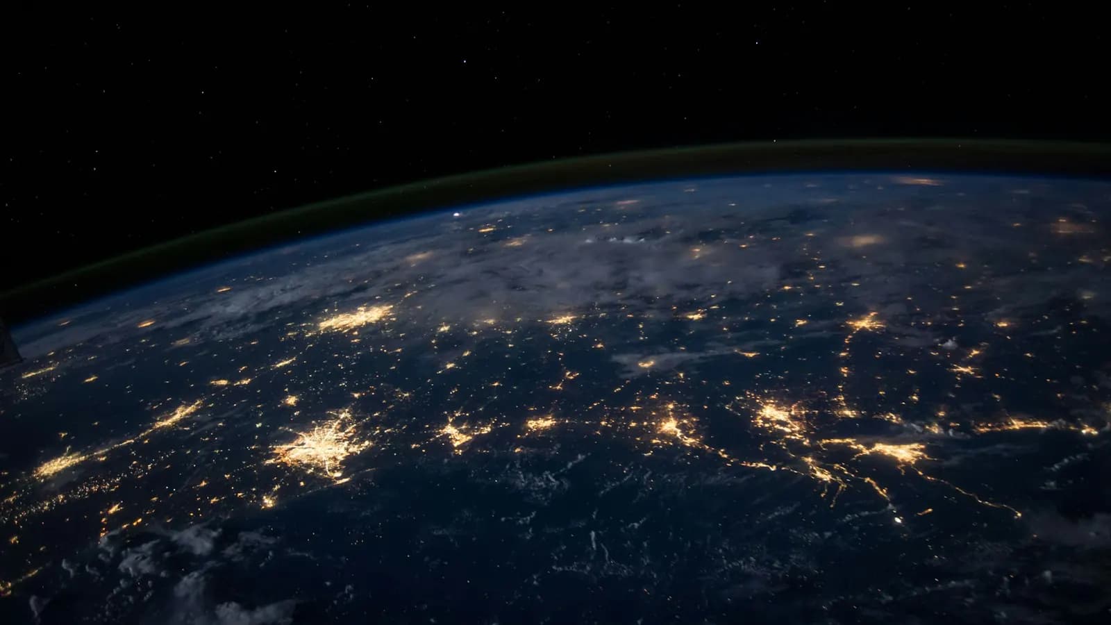 Earth at night from orbit, with city lights across the land and a thin green glow on the horizon