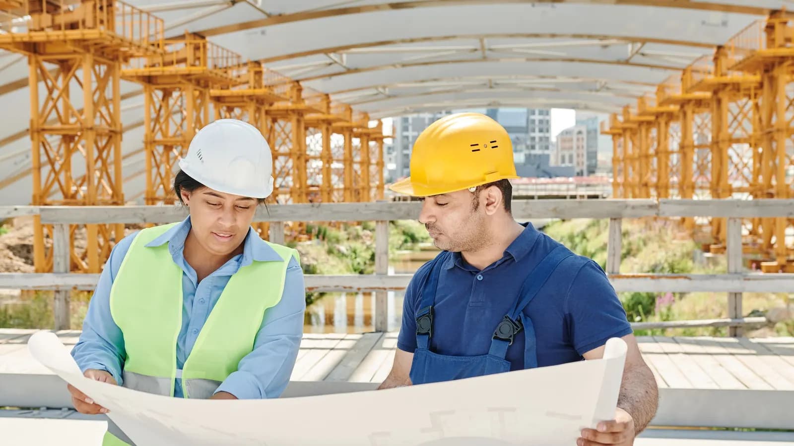 Two engineers in hard hats reviewing a large blueprint on a construction site with steel scaffolding behind them