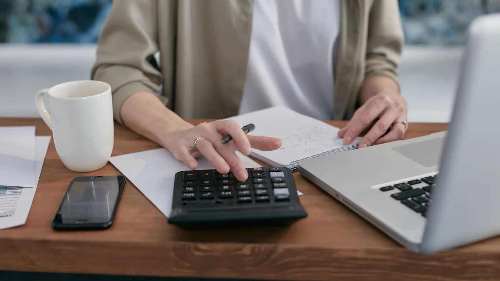 Person using a calculator at a wooden desk with a laptop, a spiral notepad and a coffee mug