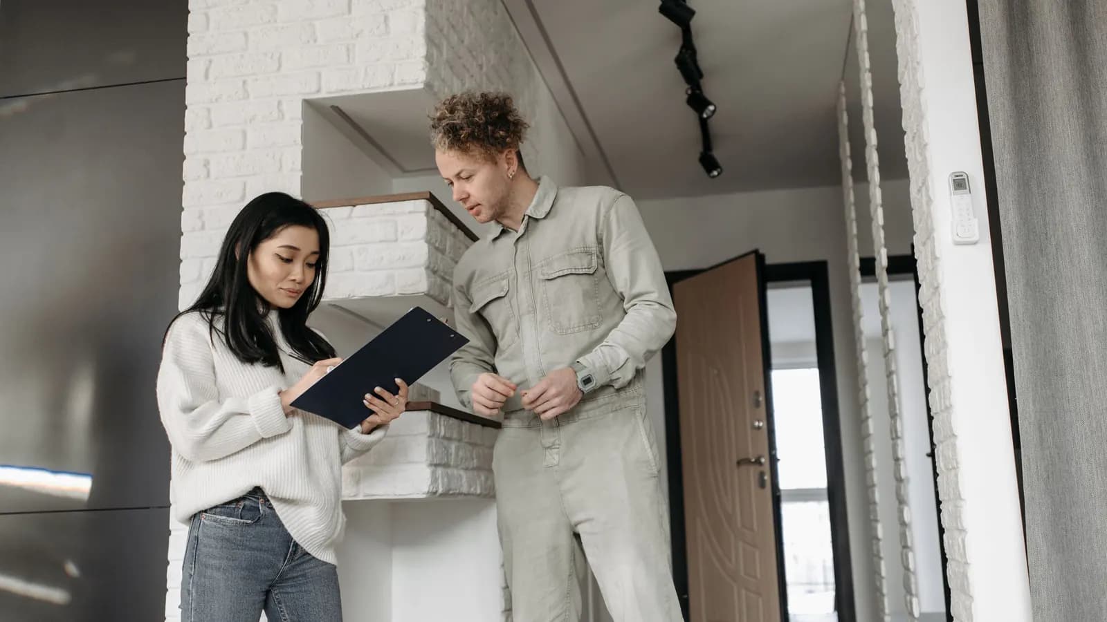 Woman in a white sweater reviewing a clipboard as a worker in gray coveralls looks on inside a home