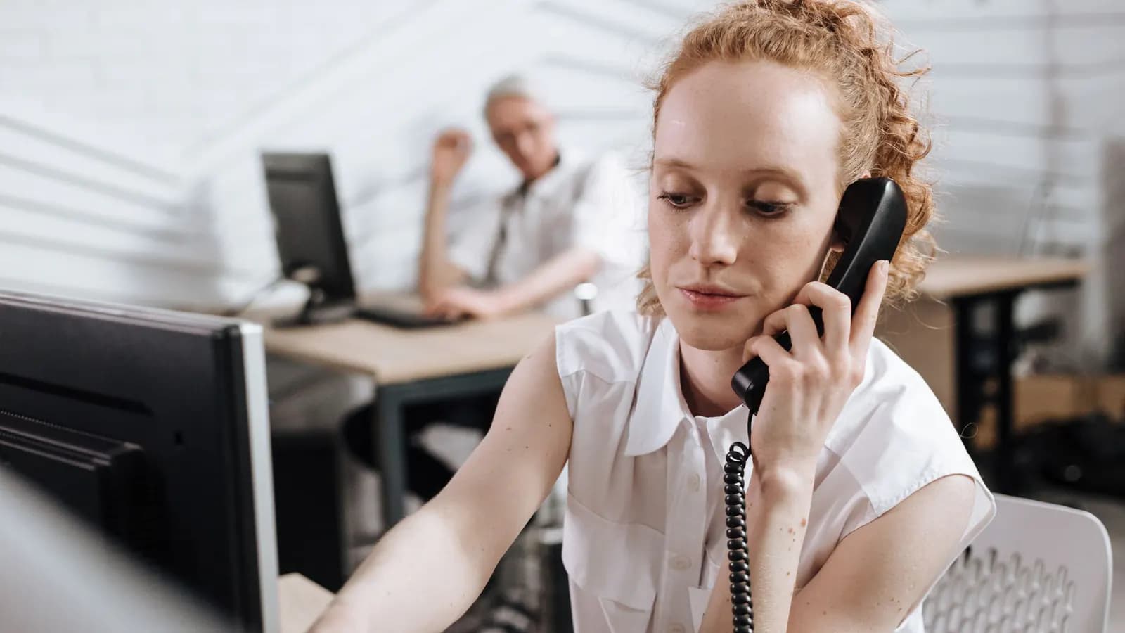 Office worker on a corded desk phone, reaching toward her computer, with a colleague at another desk behind her