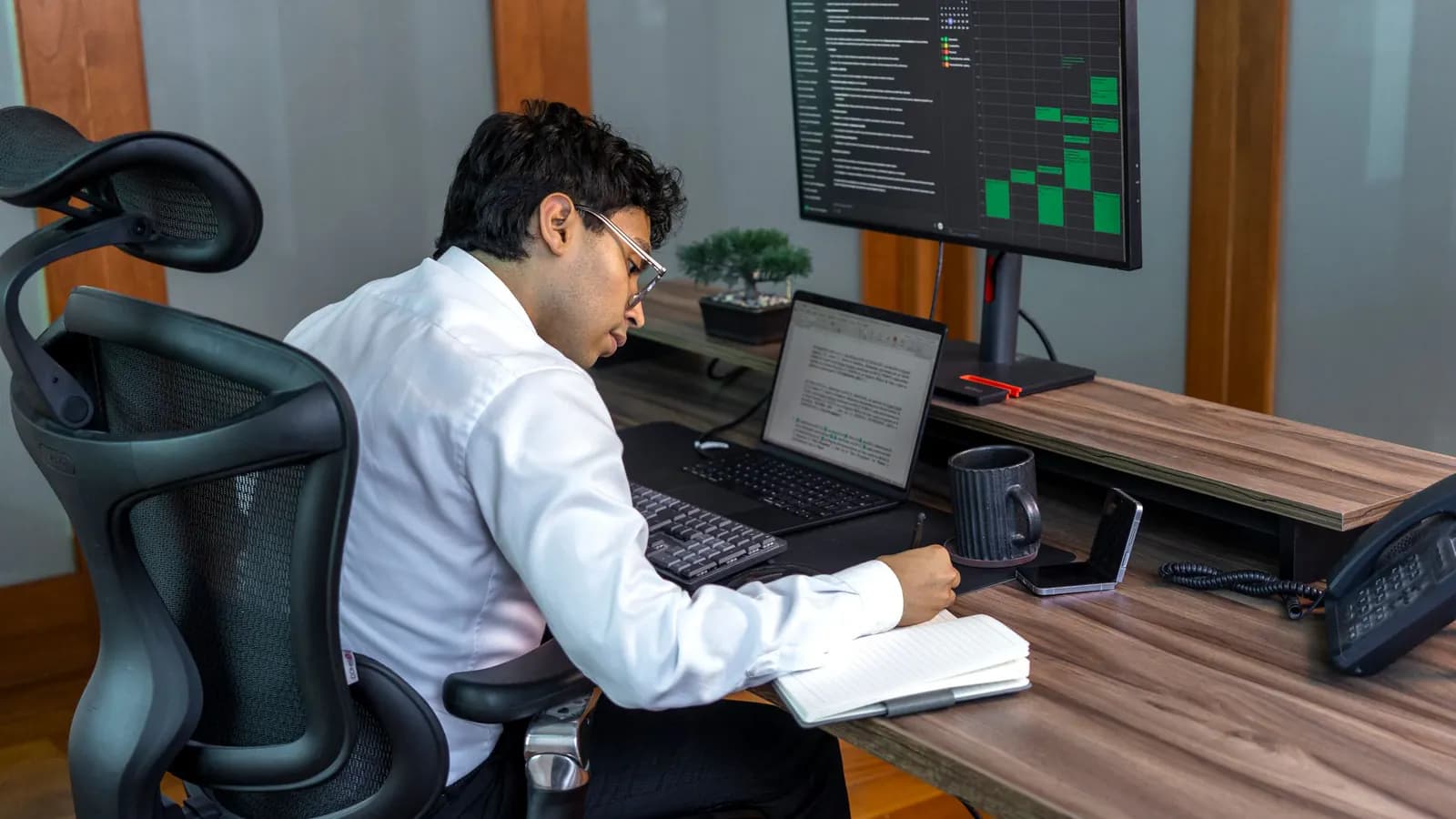 Office worker writing in a notebook beside a laptop, a desk phone and a monitor showing a calendar