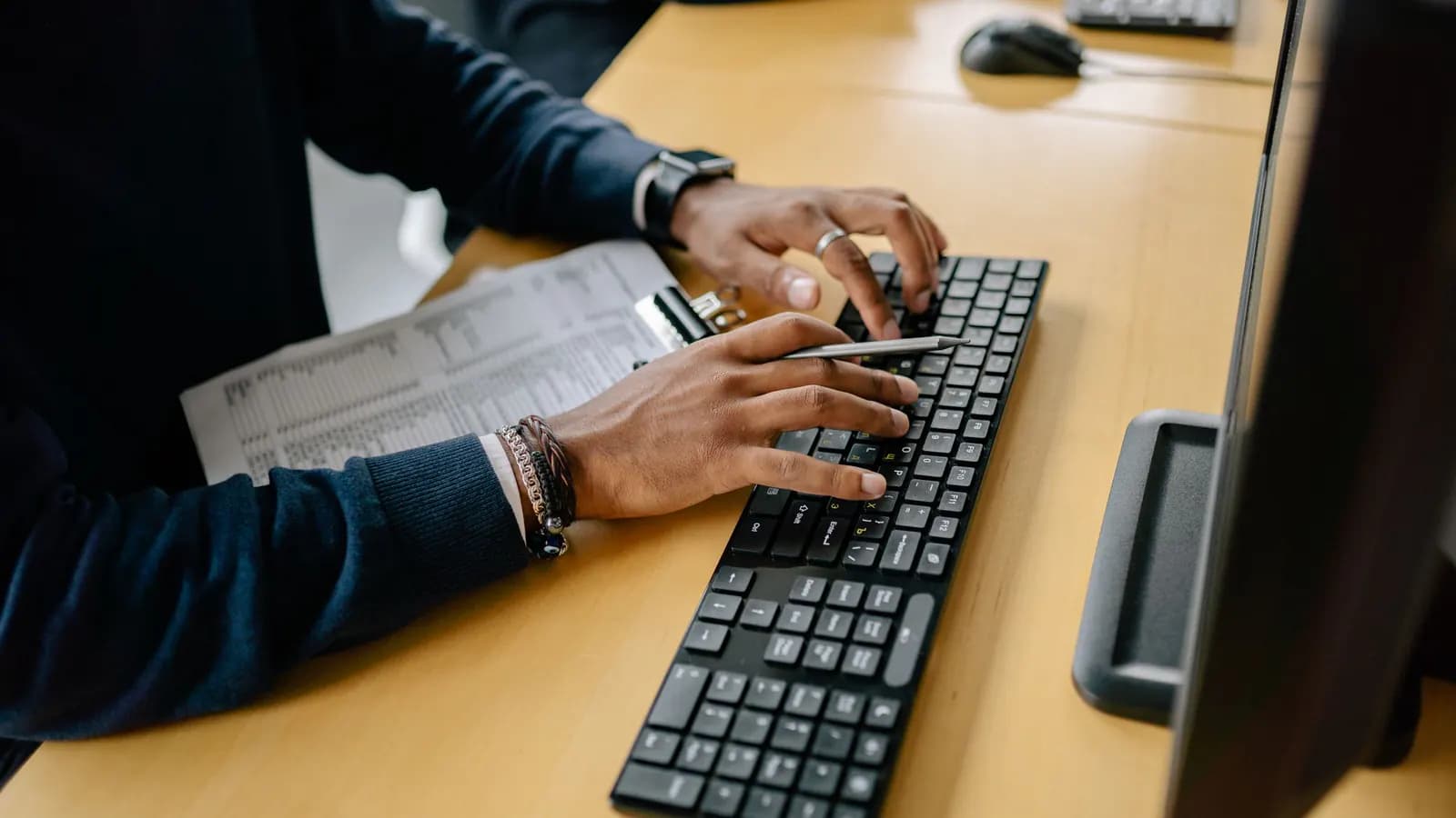 Hands typing on a keyboard at an office workstation beside a clipboard holding a printed table of figures