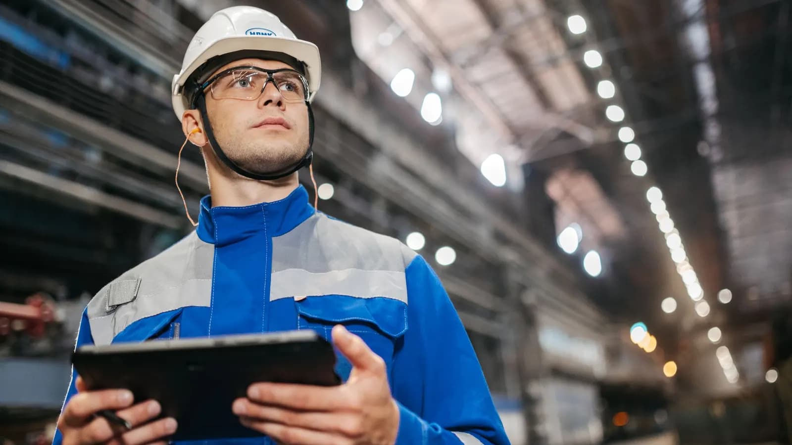 Engineer in a hard hat and safety glasses holding a tablet inside a large factory hall