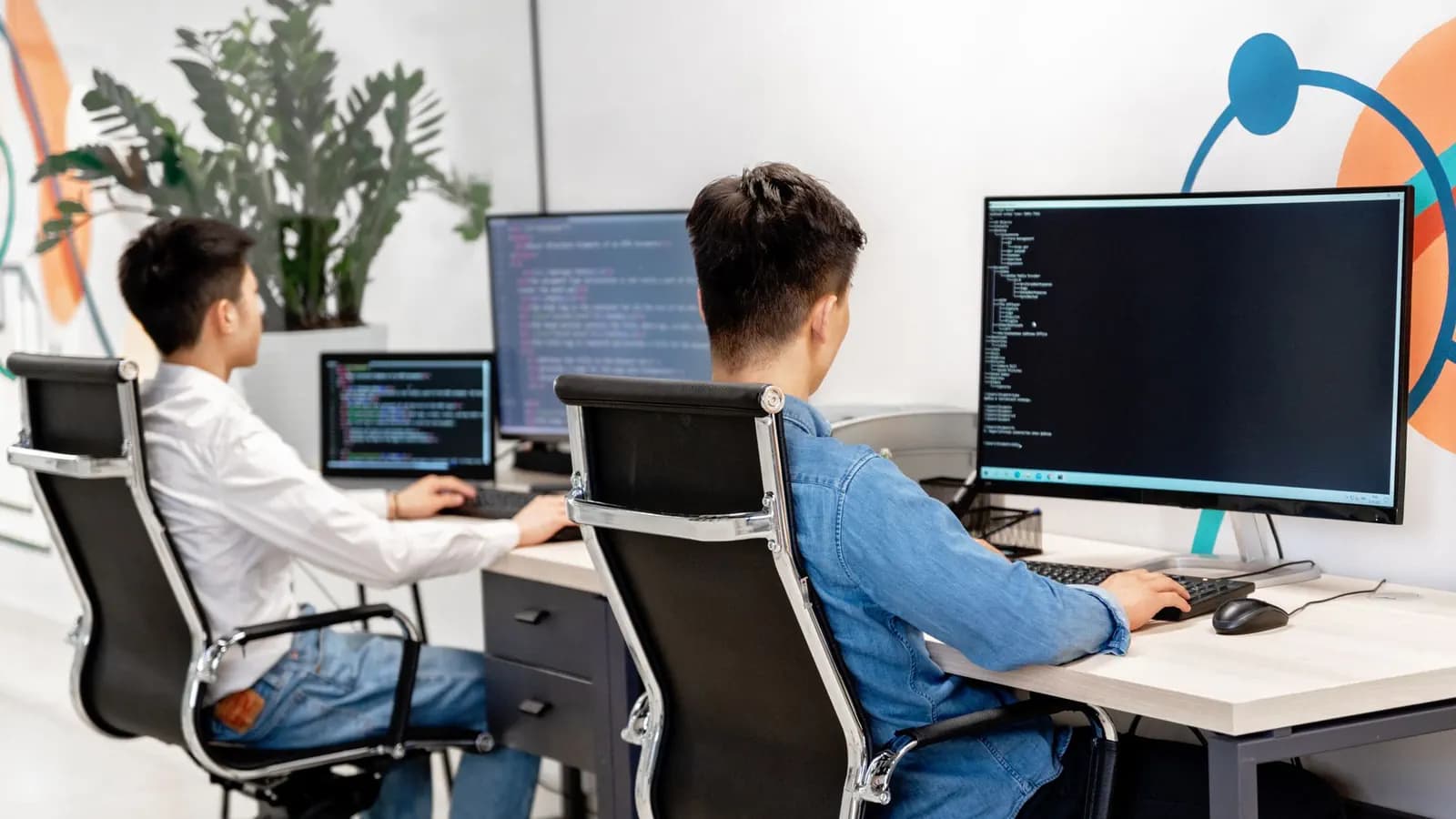 Two men at side-by-side desks working with code on large monitors in a bright office