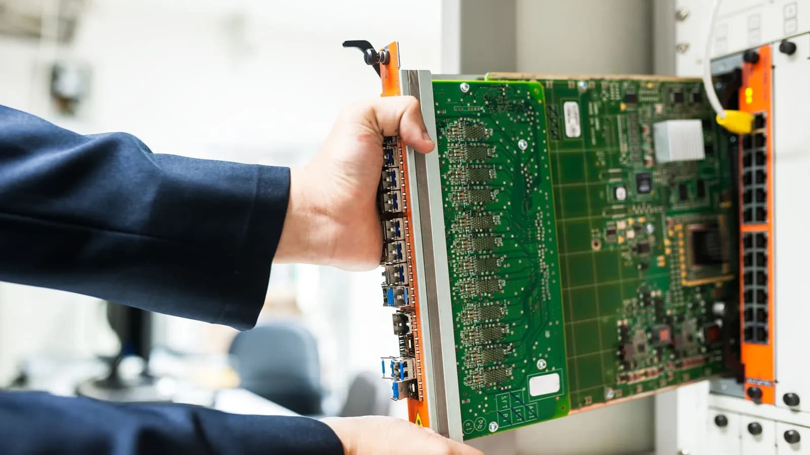Hands in navy sleeves sliding a green circuit board into a network switch chassis in an equipment room
