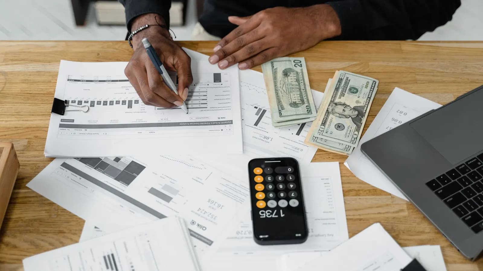 Hands writing on printed invoices next to cash, a calculator and a laptop on a wooden desk