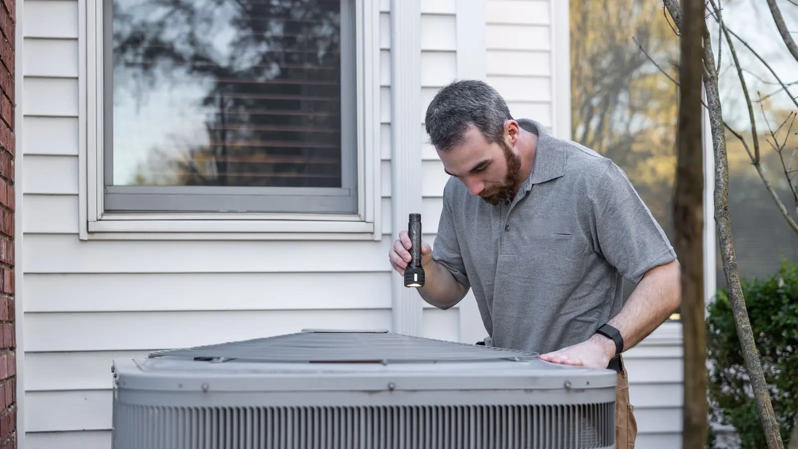Technician holding a flashlight while inspecting an outdoor air conditioning unit beside a house