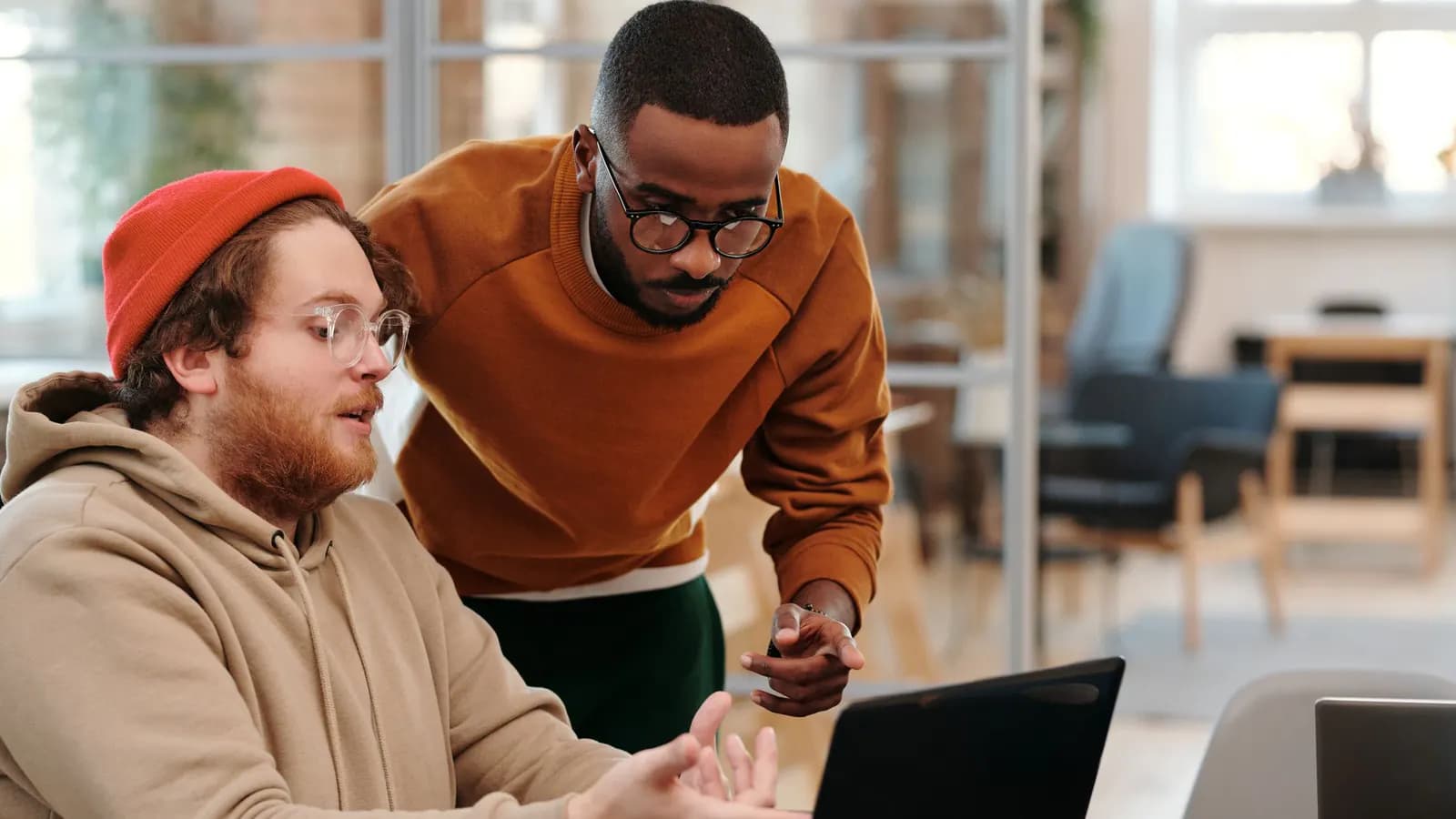 Man in a red beanie walks a colleague in an orange sweater through something on a laptop
