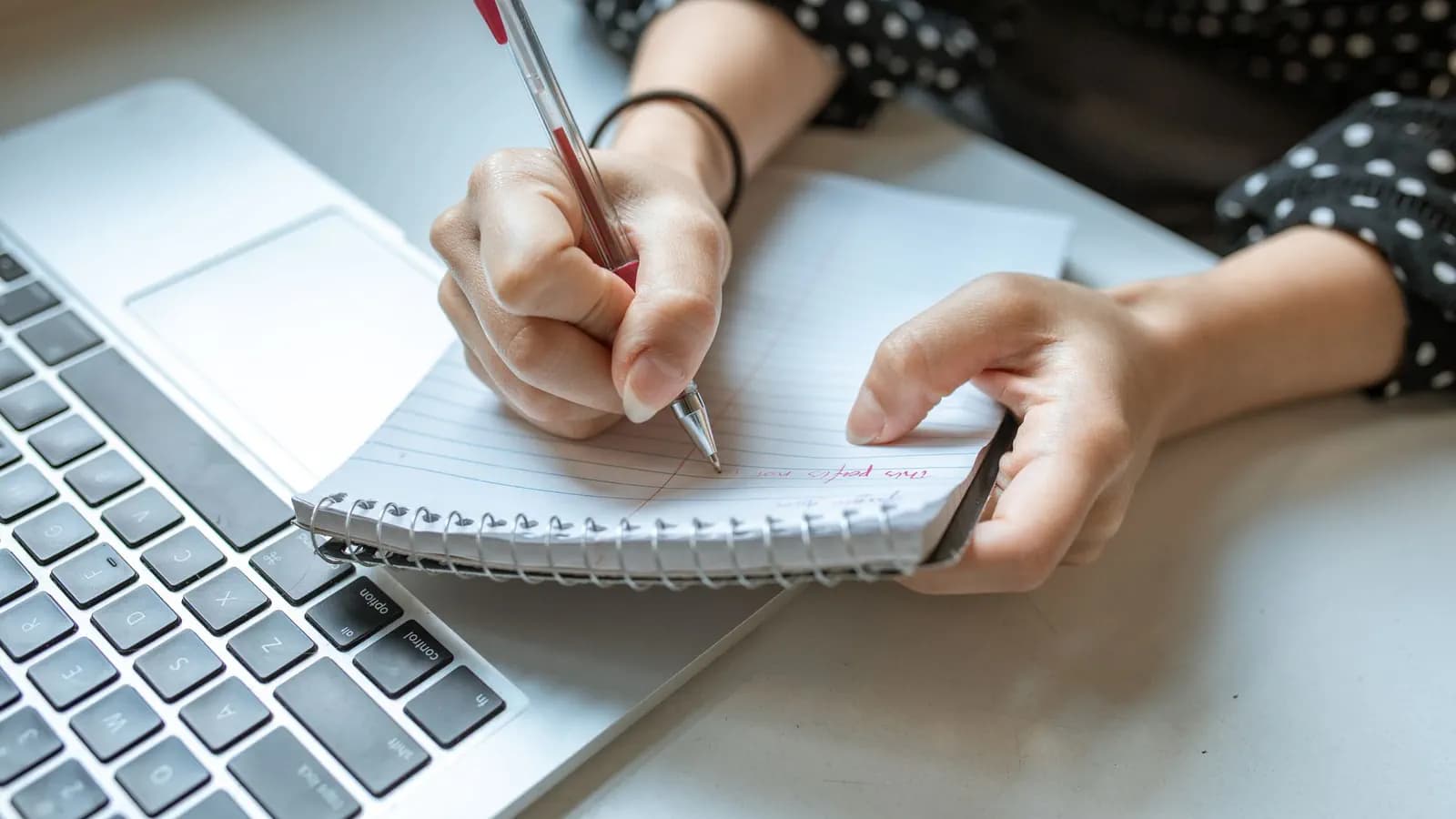 Close-up of a hand writing in a spiral notebook with a red pen beside an open laptop