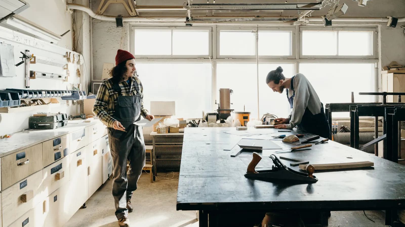 Two woodworkers in a bright shop, one carrying a sheet of paper toward the other at a long workbench