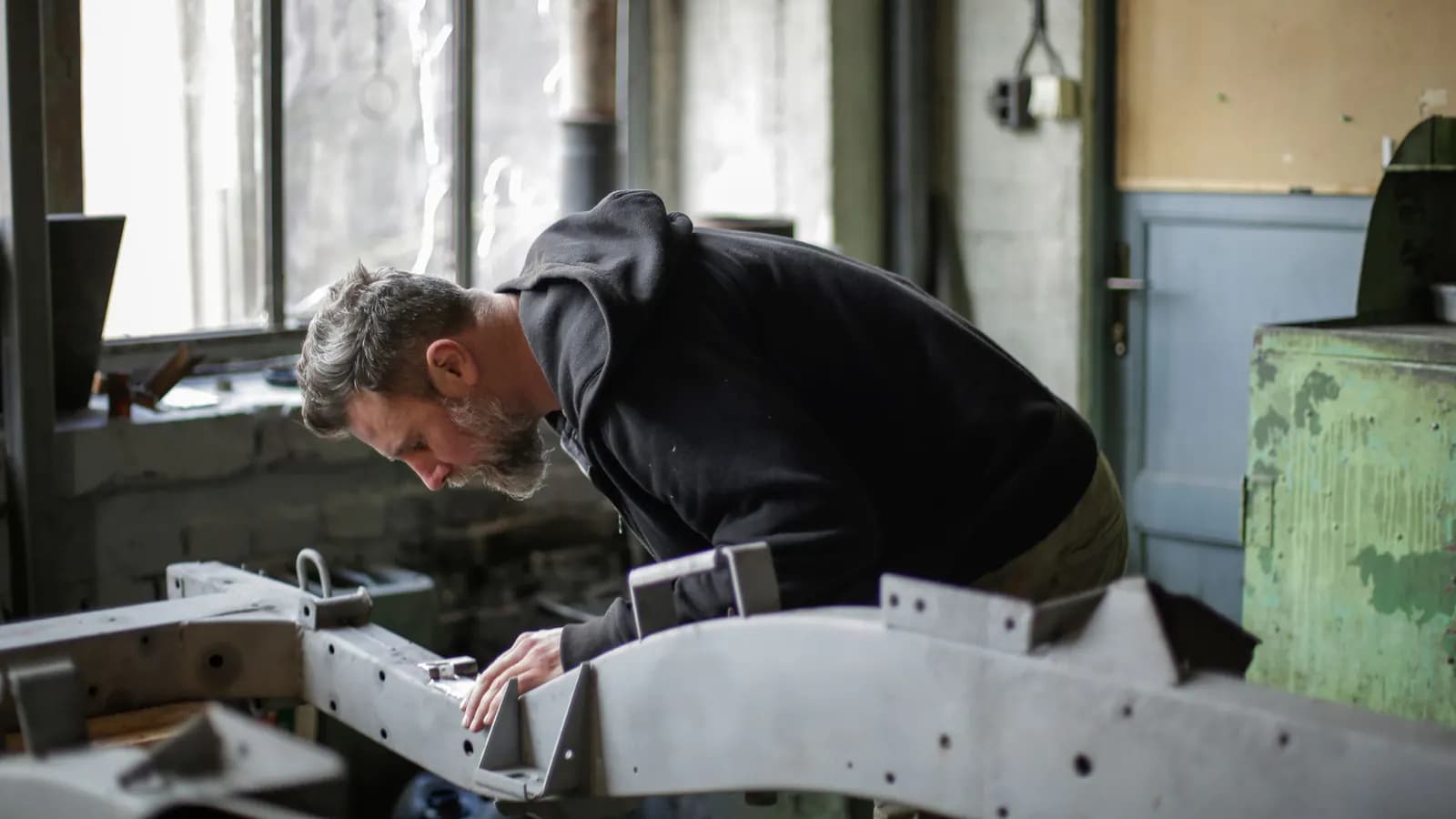 Bearded worker in a black hoodie leaning over a gray metal frame in a workshop to check it closely
