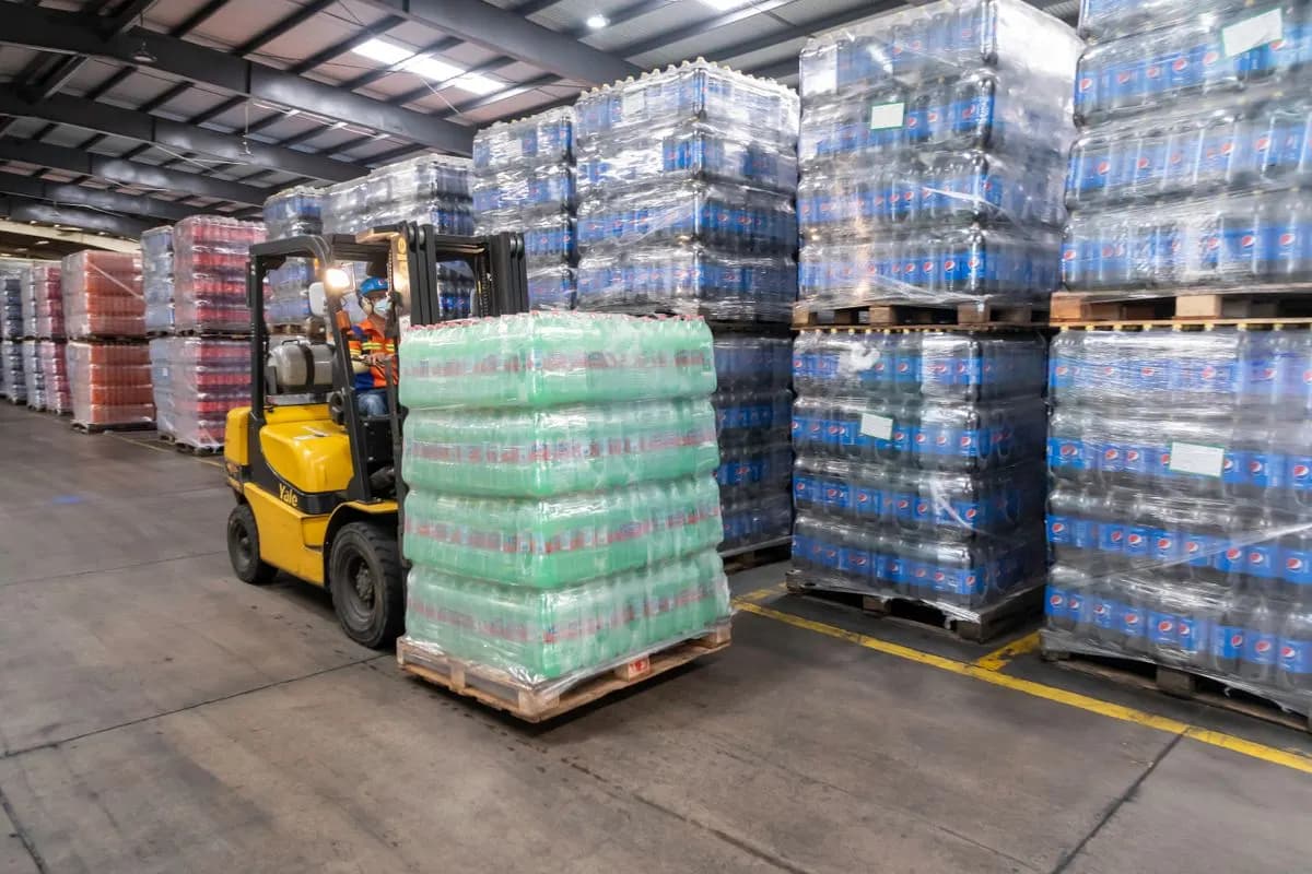 Worker driving a yellow forklift carrying shrink-wrapped bottled drinks past stacked pallets in a warehouse