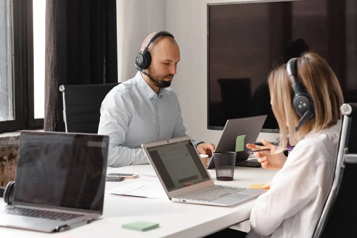 Two support agents wearing headsets work at laptops on a shared white office desk