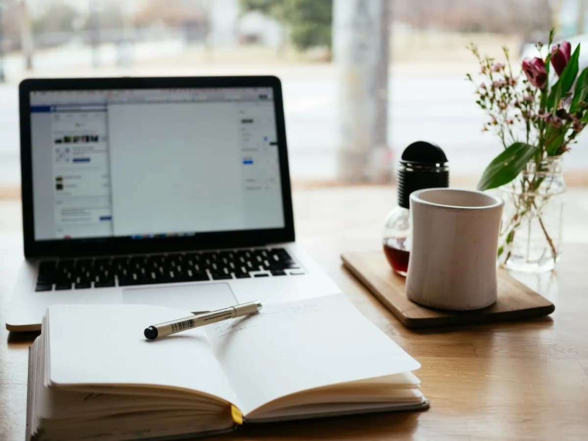 Open notebook and pen beside a laptop, a white mug and flowers on a wooden table