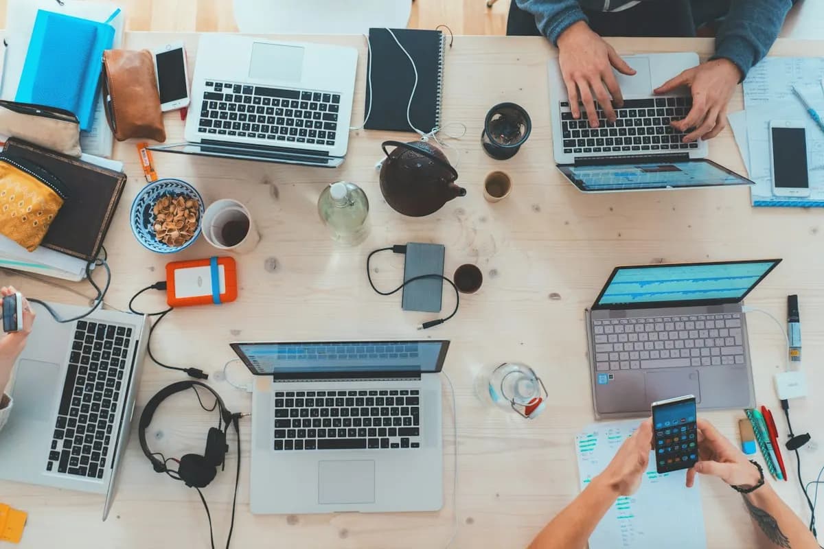 Overhead view of a shared desk with several laptops, phones, headphones, notebooks and coffee cups