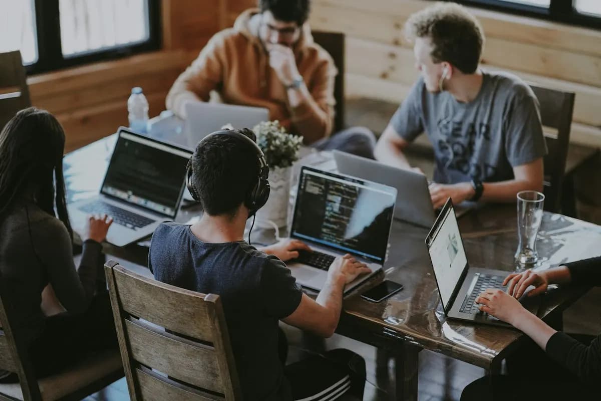 Small team of developers coding on laptops around a wooden table, one wearing headphones