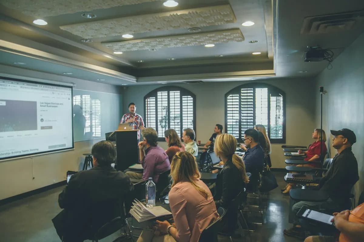 Instructor at a lectern presenting slides on a projector screen to adults seated in a classroom