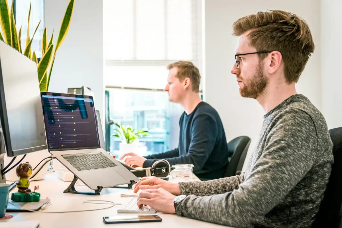 Two men at a bright office desk, one typing beside a laptop that shows code