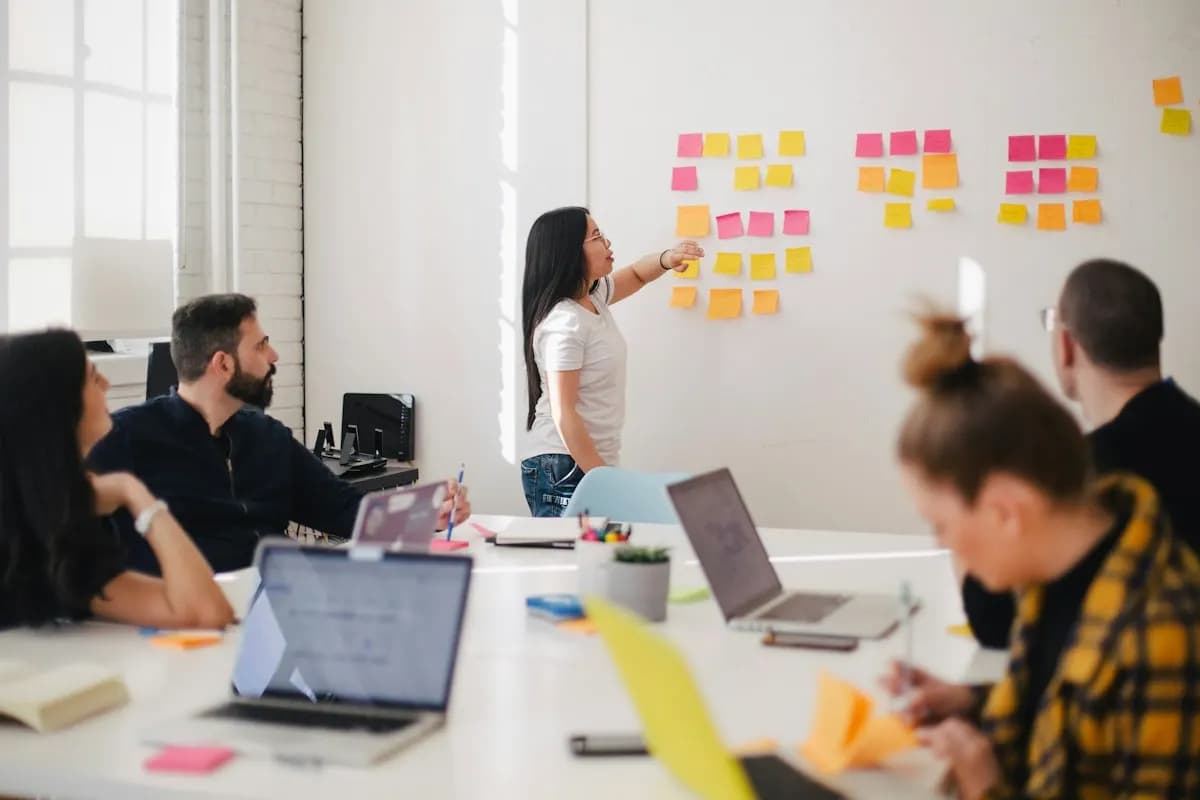 Woman points at sticky notes on a wall as colleagues with laptops watch from a table
