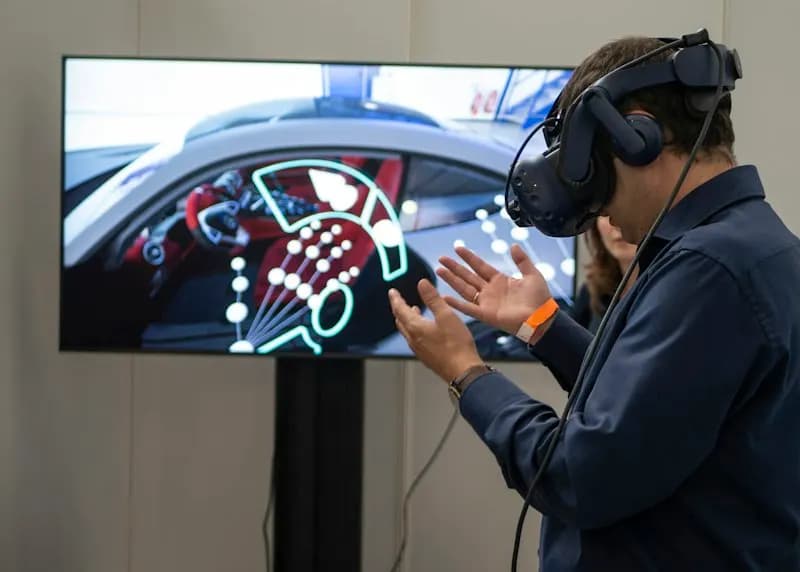 Man in a VR headset gesturing beside a screen showing a virtual car interior