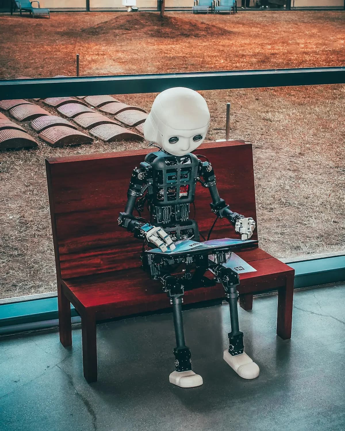 Humanoid robot with a white head sitting on a red wooden bench, reading an open book